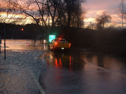 1993 Ford Bronco - A yellow pickup truck navigates through flooded streets as the sun sets, with its taillights casting reflections on the water.