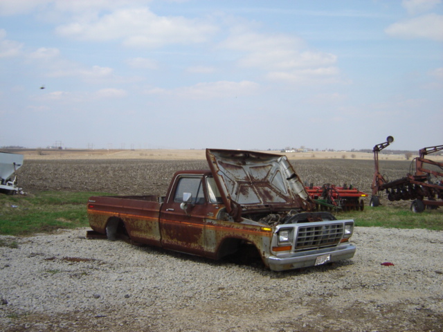 Rusty brown pickup truck with hood open, missing front wheels, parked on gravel in a field
