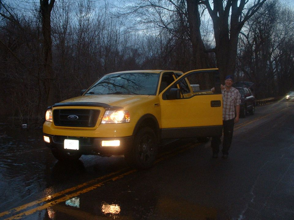 Yellow Ford F-150 pickup truck with driver's door open, parked on wet road. Another SUV is visible behind it