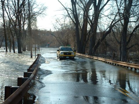 1993 Ford Bronco - A yellow Ford F-150 pickup truck navigates a flooded road, with trees lining the water-covered asphalt.