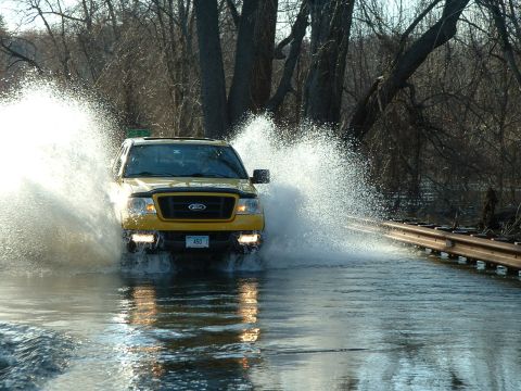 1993 Ford Bronco - A yellow Ford F-150 truck splashes through a flooded road, kicking up water on both sides.