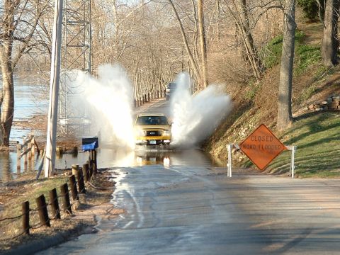 1993 Ford Bronco - A yellow SUV drives through a flooded road, kicking up water, past a "CLOSED ROAD FLOODED" sign.