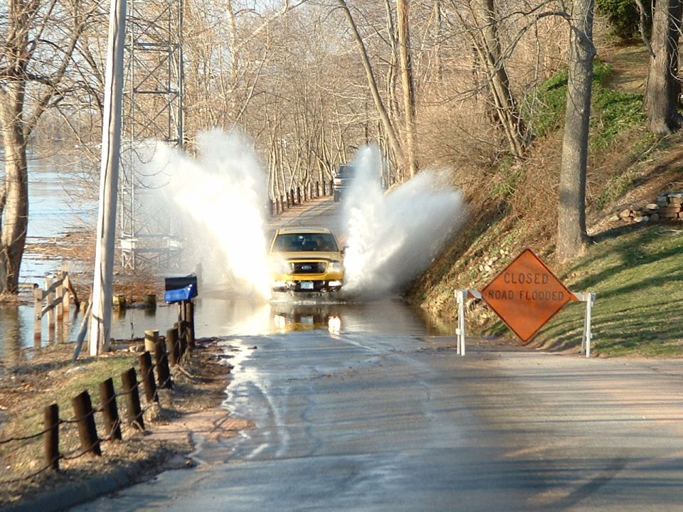 Yellow SUV driving through flooded road, creating large splashes. A "CLOSED ROAD FLOODED" sign is visible