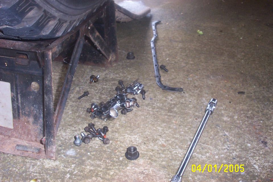 Engine intake manifold and bolts scattered on concrete next to a tire on a jack stand