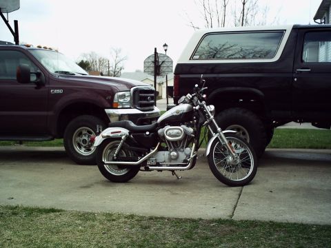 1986 Ford Bronco - A motorcycle is parked between a maroon Ford F-250 and a black Ford Bronco, with a basketball hoop in the background.