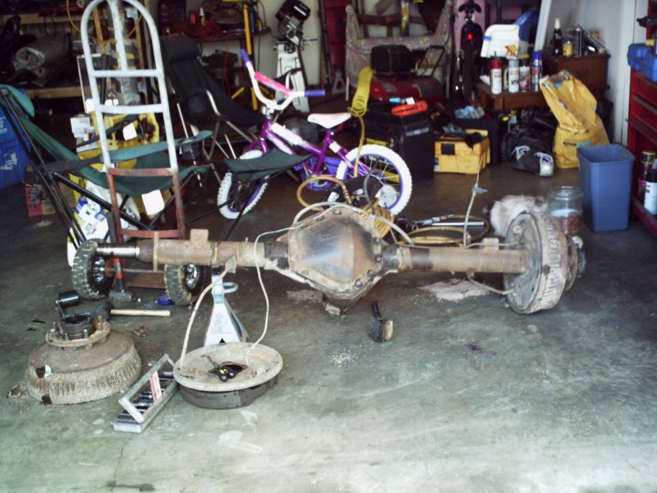 Rear axle assembly with brake drums and hubs disassembled, resting on jack stands in a garage