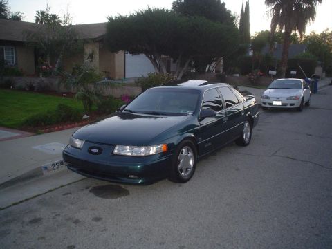 1993 Ford Taurus - A dark green Ford Taurus is parked on a residential street next to another white car.