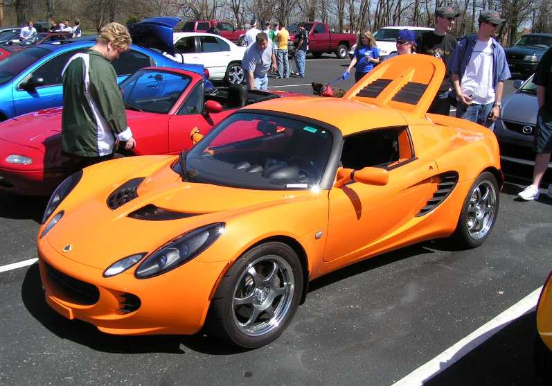 Orange Lotus Elise roadster at a car show with spectators and red roadsters nearby