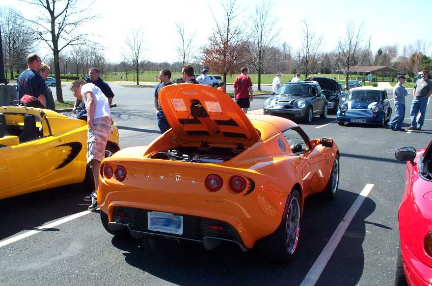 Orange Lotus Elise with engine cover raised at a car meet, yellow Elise and Minis nearby