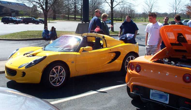 Yellow Lotus Elise roadster at a car meet beside an orange Elise with rear clamshell open