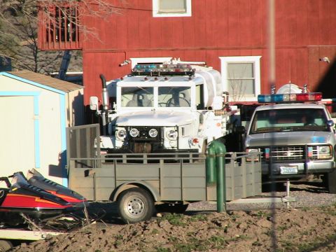 1978 Ford Bronco - A white military truck with a water tank and emergency lights is parked next to a silver pickup truck, with a snowmobile on a trailer in th…