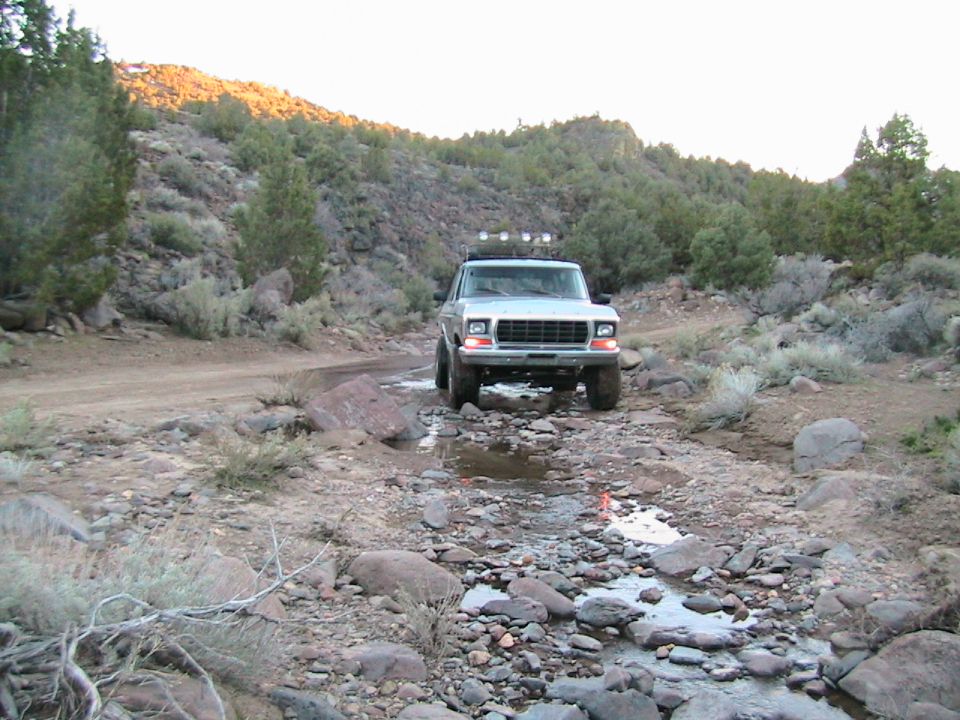 1978 Ford Bronco driving through a rocky stream bed with scrub brush and hills in background