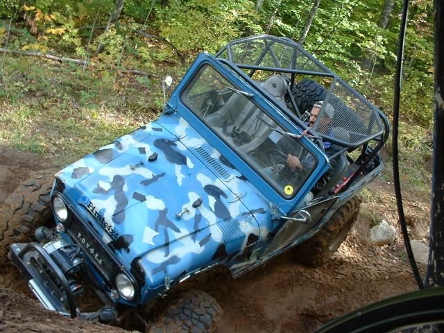 Blue camo open-top Toyota 4x4 with roll cage and winch off-roading in mud on a forest trail