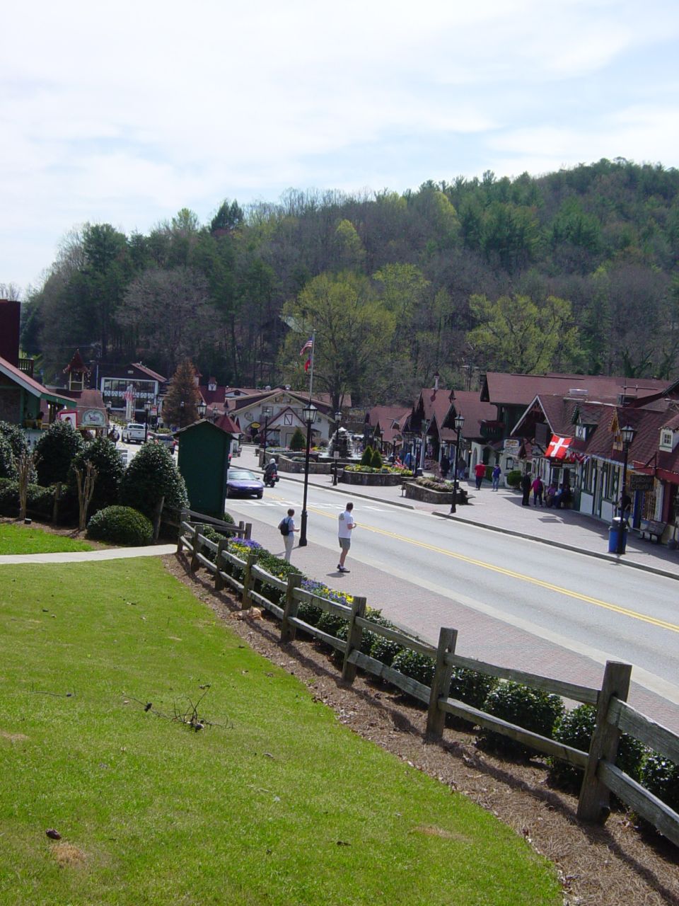 Street scene in a town with Bavarian-style architecture, trees on a hill, and a wooden fence in the foreground