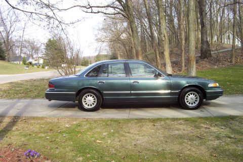 1996 Ford Crown Victoria - A green 1996 Ford Crown Victoria sedan is parked on a driveway, with trees and houses in the background.