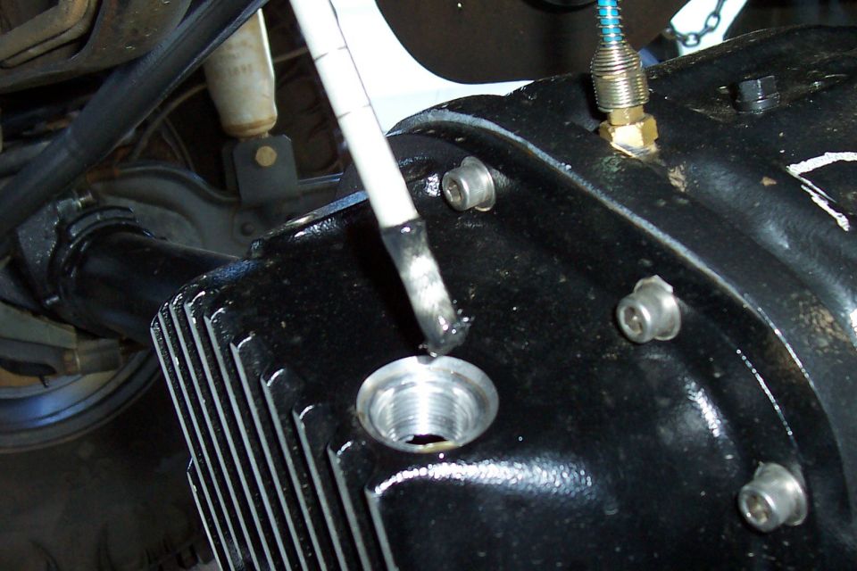Close-up of a black differential housing with a tool inserting into a fill hole. A shock absorber is visible in the background