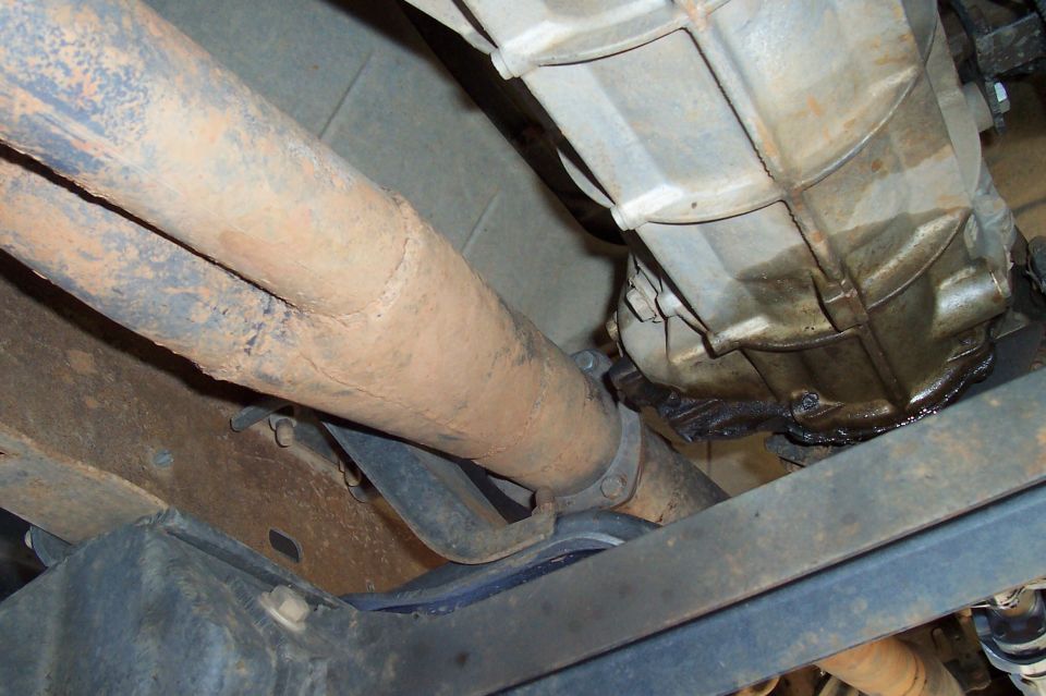 Rusty exhaust downpipe routed alongside an oily aluminum transmission case under a truck