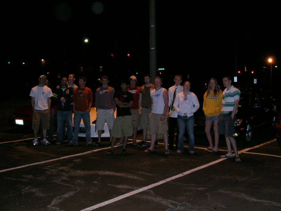 Group of people in a parking lot at night with cars