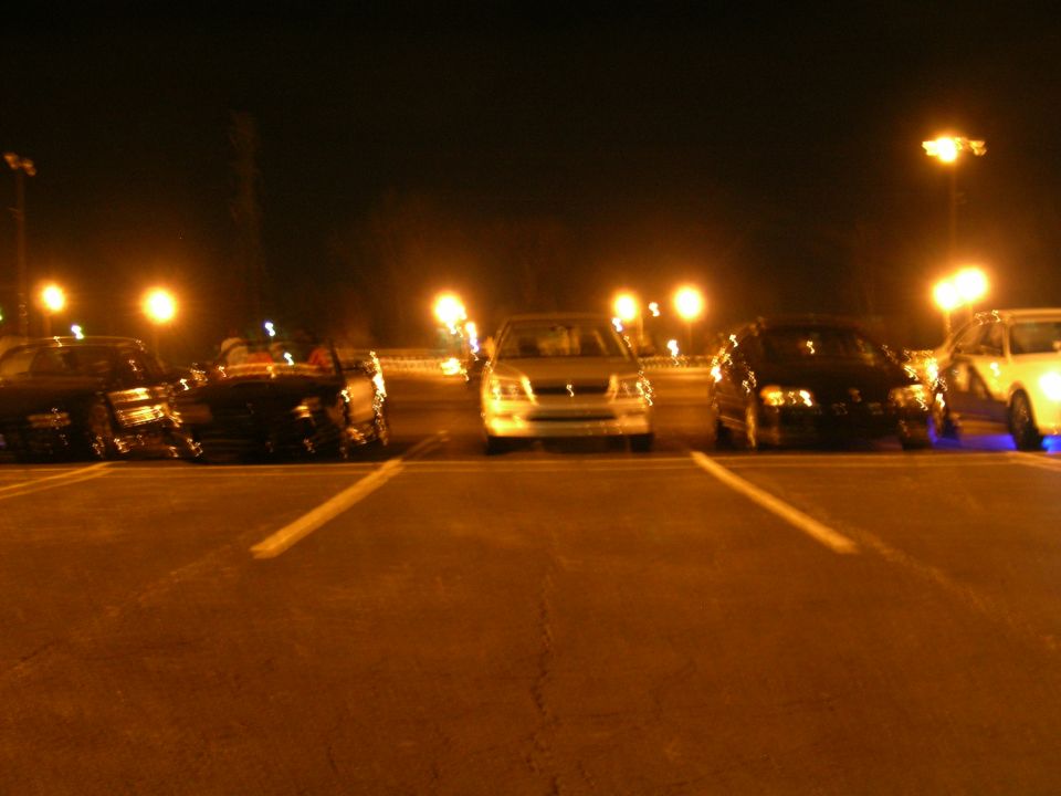 Several cars parked in a lot at night, illuminated by streetlights