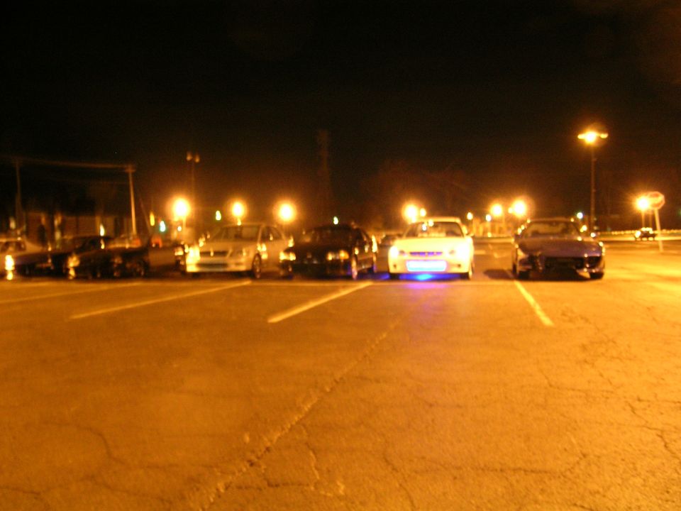 Several cars parked in a lot at night, one with blue underglow