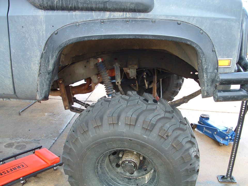 Lifted gray pickup's front wheel well with big off-road tire, leaf springs, floor jack and creeper nearby