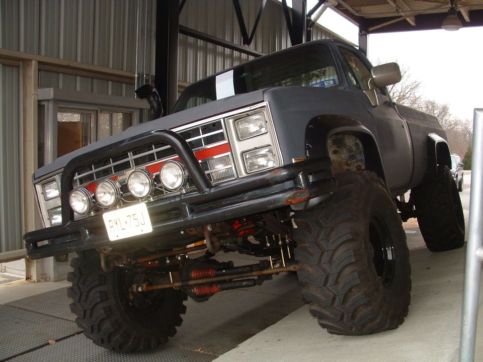 Lifted gray square-body Chevy pickup with brush guard, off-road lights and big mud tires under a carport.