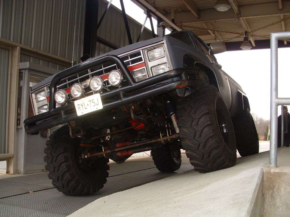 Lifted black Chevy pickup, low front angle, big mud tires, brush guard with round lights