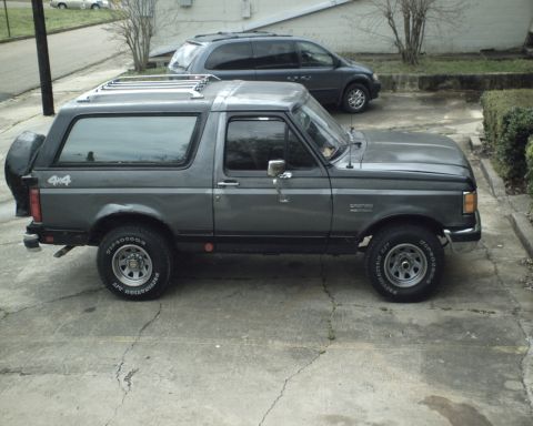 1987 Ford Bronco - A gray 1987 Ford Bronco is parked on a concrete driveway next to a dark minivan.