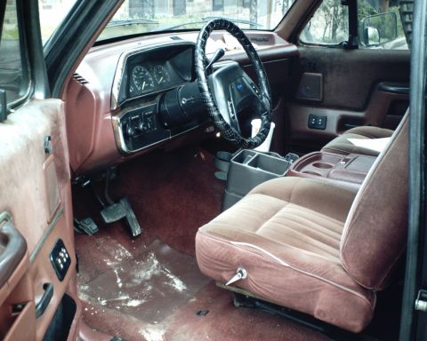 1987 Ford Bronco - The interior of a 1987 Ford Bronco shows brown velour seats and dashboard, with a water stain on the floor mat.