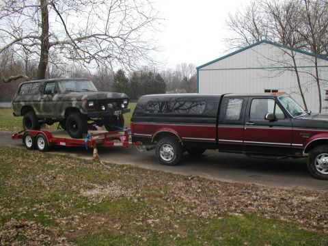 1979 Ford Bronco - A camouflage Ford Bronco is transported on a trailer, towed by a two-tone pickup truck.