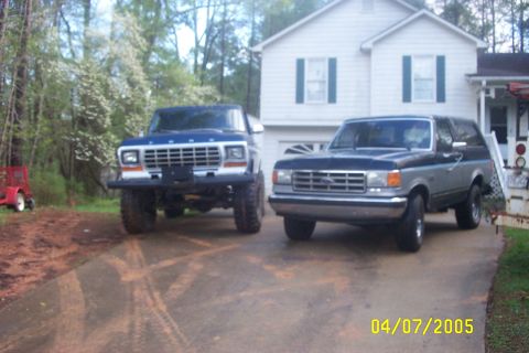 1979 Ford F-150 - Two Ford trucks are parked in front of a house, one blue and white, the other gray and black.