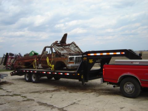 1992 Ford Bronco - A heavily damaged and rusted pickup truck, appearing to be from the early 1980s, is being transported on a flatbed trailer.