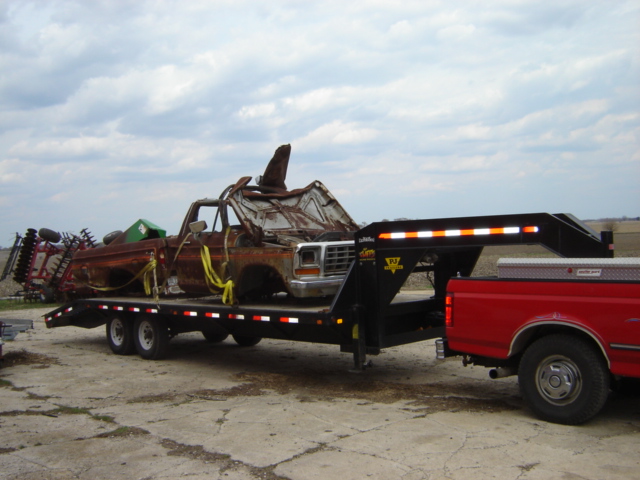 Rusted 1980s pickup truck on a trailer, front 3/4 view. Another vehicle is visible on the trailer
