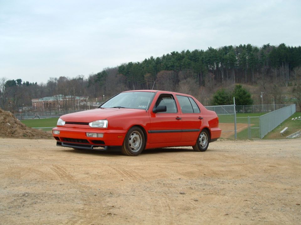 Red 1994 Volkswagen Jetta sedan parked on dirt lot with trees and a baseball field in background
