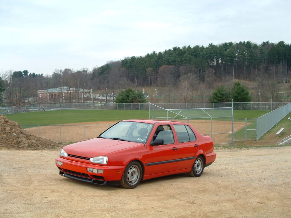 Red sedan parked on dirt lot, baseball field in background