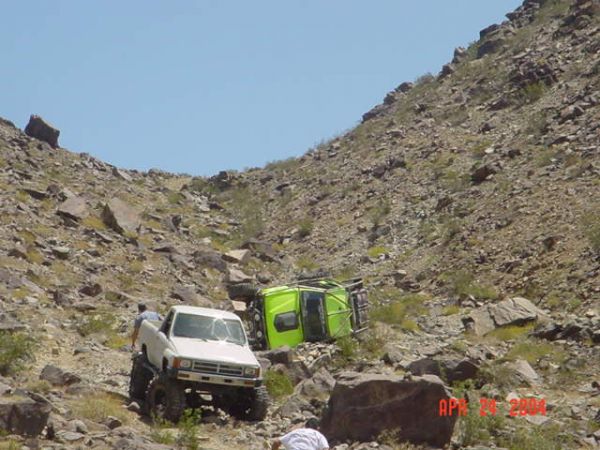 White off-road pickup on rocky desert hill beside a green rolled-over vehicle