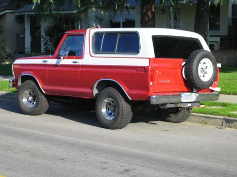 1990 Ford Bronco - Rear three-quarter view of a red-and-white Ford Bronco with a tailgate-mounted spare tire and chrome wheels parked at a residential curb.