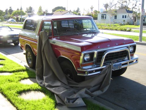 1990 Ford Bronco - red Ford Bronco with chrome grille and FORD hood lettering, a fabric cover partly slipping off the front, parked on a residential street.