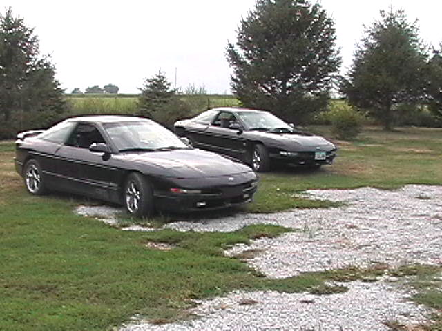 Two dark Ford Probe coupes parked on grass in a rural yard with trees behind