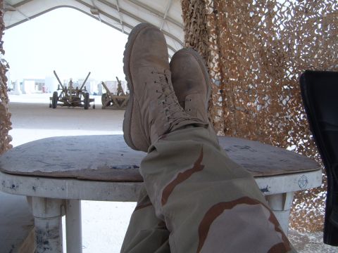 1988 Ford Bronco - A desert scene artillery pieces and camouflage netting, with a person's boots and camouflage pants in the foreground.