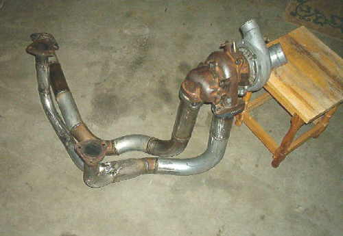 Turbocharger attached to a rusty exhaust manifold with two pipes, resting on a concrete floor next to a wooden stool