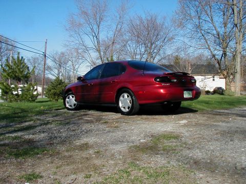 1997 Ford Taurus - Red 1997 Ford Taurus sedan seen from the rear, parked on a gravel lot with trees and a house visible.