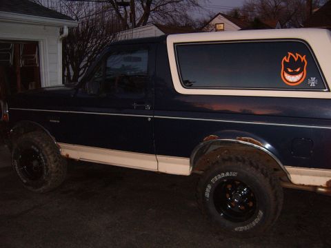 1993 Ford Bronco - Dusk side profile of a lifted dark blue full-size Ford Bronco with a white removable hardtop, parked in a driveway. It sits on black steel…