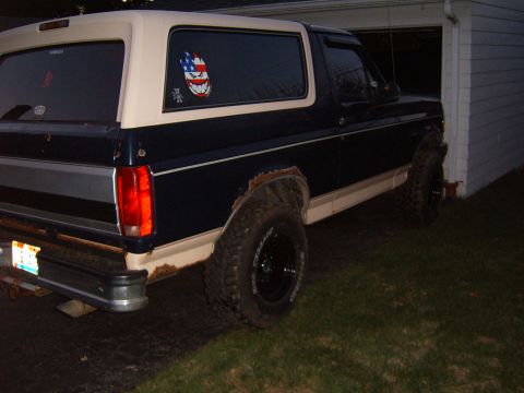 1993 Ford Bronco - Night-time three-quarter rear view of a lifted dark blue and white full-size Ford Bronco parked on grass beside a white garage. It rides on…