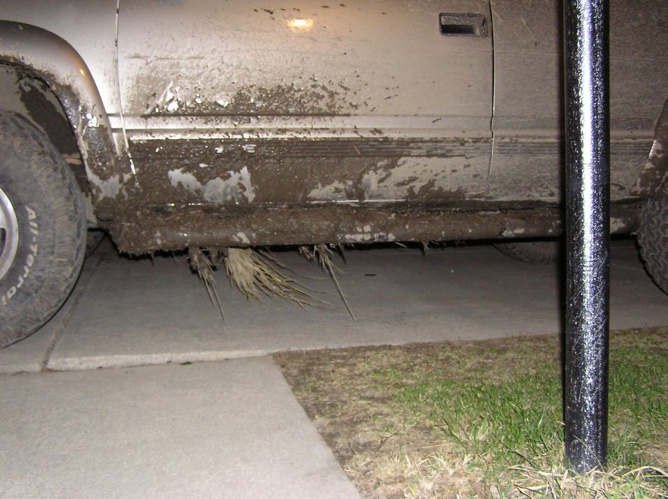 Mud covers the side panel and lower body of a truck, with vegetation hanging from the undercarriage. A tire is visible on the left