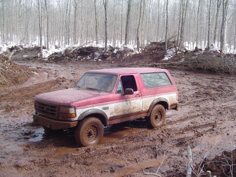 2000 Chevrolet Tahoe - A red and white Ford Bronco navigates a muddy terrain with trees and snow in the background.