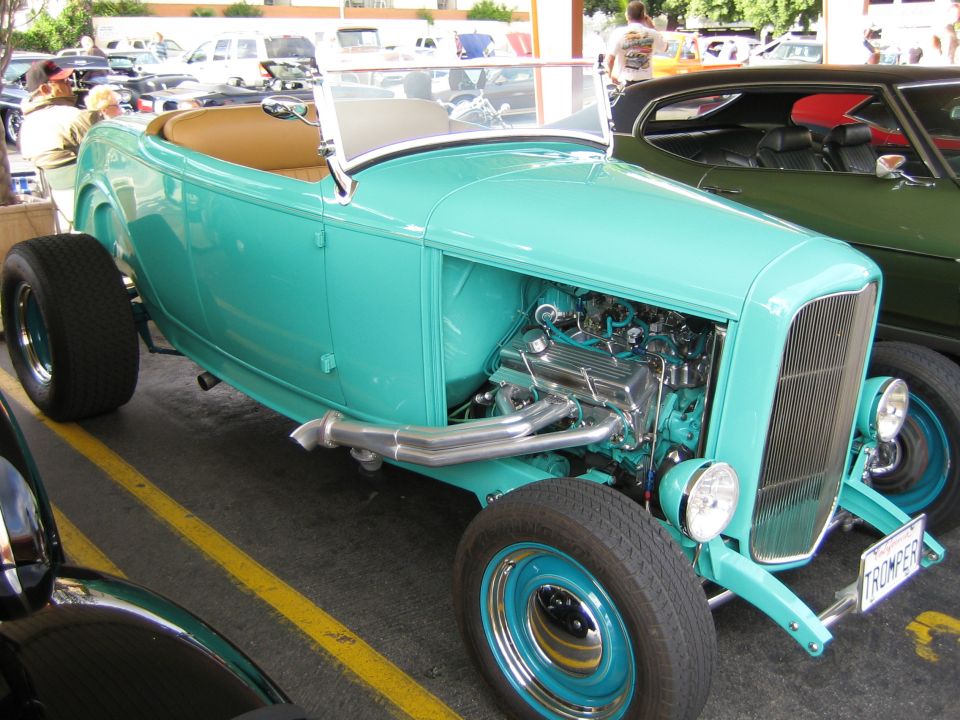 Turquoise hot rod with exposed engine and chrome exhaust pipes, parked next to a dark green car