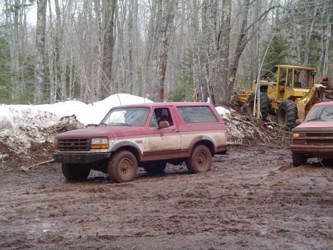 2000 Chevrolet Tahoe - A red and white Ford Bronco navigates a muddy terrain, with construction equipment and another vehicle visible in the background.