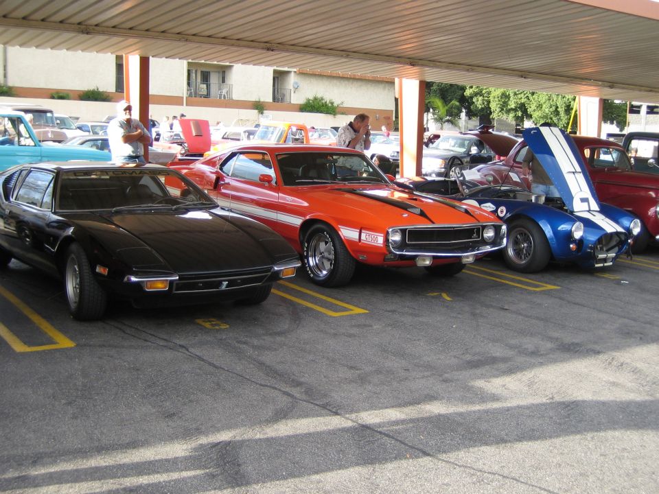 Black De Tomaso Pantera, orange muscle car, and blue Cobra parked in a lot
