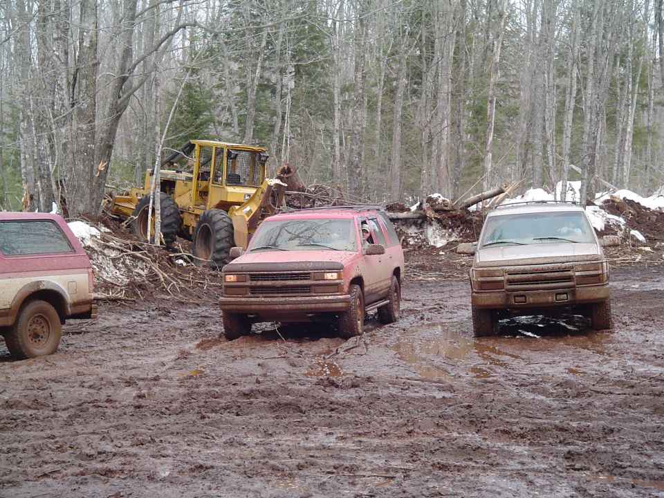 Red Chevrolet Tahoe driving through mud, front view. Other vehicles and a loader are visible in the background